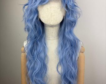 May include: A light blue wig with long, wavy hair and bangs. The wig is displayed on a mannequin head with a neutral-colored face and wooden arms. The hair has a natural, flowing appearance.