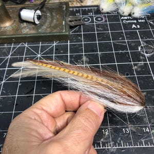 May include: A handmade fly fishing lure with brown, white, and yellow feathers. The lure is held in a hand against a black cutting mat with other fly tying materials in the background.