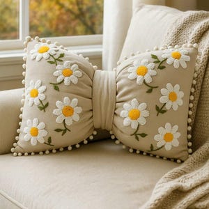 Beige linen bow pillow with handmade daisy embroidery, styled on a modern minimalist sofa.