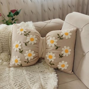 Beige linen bow pillow with handmade daisy embroidery, styled on a modern minimalist sofa.