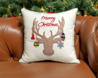 May include: A square white throw pillow featuring a textured tan embroidered reindeer head with large antlers. The antlers are decorated with small hanging ornaments including a red hat, a green and yellow bauble, and a silver star. The text Merry Christmas is embroidered in red above the reindeer.