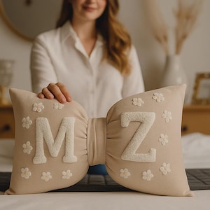Peut inclure: Un coussin beige en forme de nœud papillon avec les lettres "MZ" en blanc, orné de petites fleurs blanches. Le coussin est posé sur un lit, avec une personne en arrière-plan.