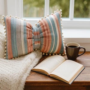 Peut inclure: Un coussin en forme de nœud à rayures verticales pêche, bleu et blanc, bordé de pompons blancs. Une tasse de café marron et un livre ouvert reposent sur une surface en bois près d'une fenêtre.