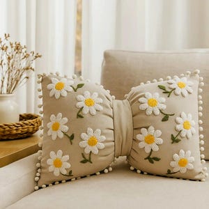 Beige linen bow pillow with handmade daisy embroidery, styled on a modern minimalist sofa.