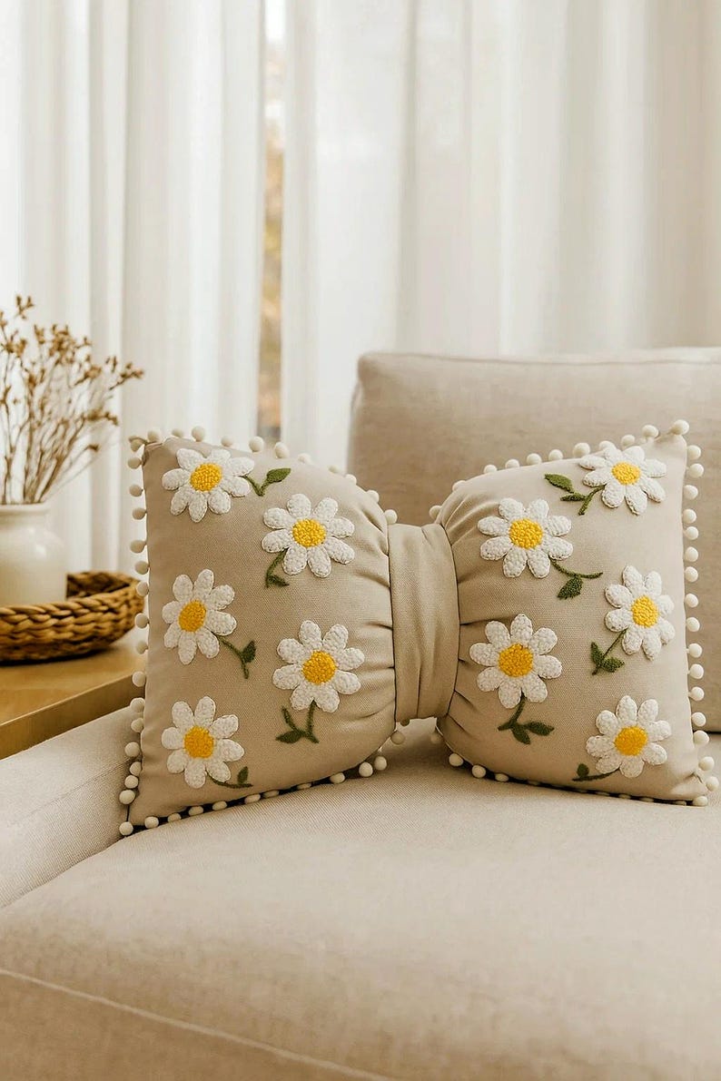 Beige linen bow pillow with handmade daisy embroidery, styled on a modern minimalist sofa.