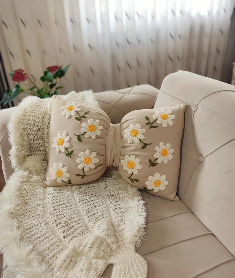 Beige linen bow pillow with handmade daisy embroidery, styled on a modern minimalist sofa.