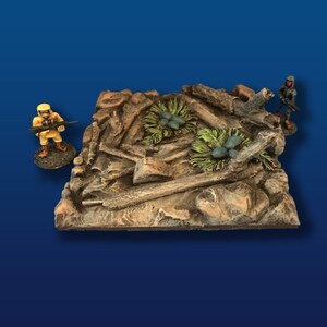 May include: A miniature terrain piece for tabletop gaming, featuring a rocky, brown surface with scattered pieces of wood and two small green bushes with blue berries. Two miniature figures are standing on the terrain, one in yellow and one in blue.