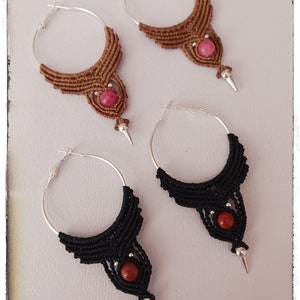 May include: A pair of handmade earrings with a geometric design. The earrings are made with brown and black cord and feature a pink and red stone accent. The earrings are attached to silver hoops.