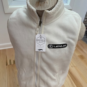 May include: Cream-colored fleece vest with a full zipper and a high collar. The vest features a black Lexus logo patch and a tag that reads "UPCYCLE GARAGE - APPAREL FOR VINTAGE CAR LOVERS". The vest is displayed on a mannequin.