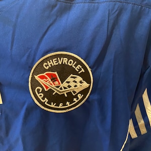 May include: A close-up of a blue jacket featuring a Chevrolet Corvette logo patch. The patch is circular with the word "Chevrolet" at the top and "Corvette" at the bottom. The logo includes a checkered flag and a red flag.