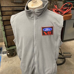 May include: A gray fleece vest with a full-length zipper and a high collar. The vest features a Ford V8 patch on the chest. The vest is displayed on a mannequin.