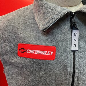 May include: Gray fleece jacket with a black zipper and a red Chevrolet logo patch. The jacket has a collared neckline and a tag that says "USA".