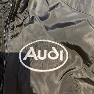 May include: Black jacket with a white Audi logo patch. The jacket has a zipper closure and a quilted design. The Audi logo is in a white oval with a black background. The jacket appears to be made of a nylon material.