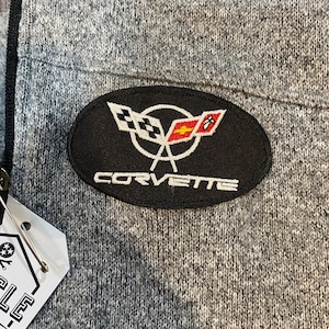 May include: Gray zip-up jacket with a black oval patch featuring the Corvette logo. The patch displays crossed flags and the word "CORVETTE" in white. The jacket has a textured knit fabric and a black zipper.