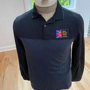 May include: A long-sleeved black polo shirt with a blue striped design across the chest. The shirt features a small patch with the Union Jack flag and the text "John Player Team Lotus".