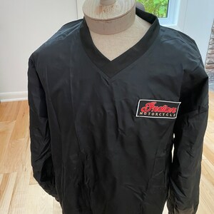 May include: Black pullover jacket with a V-neck collar. The jacket features a rectangular patch with the words "Indian MOTORCYCLE" in red and white. The jacket is displayed on a mannequin.