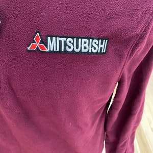 May include: A burgundy fleece jacket featuring a patch with the Mitsubishi logo and text. The patch is rectangular with a black border, white text, and a red and white logo. The jacket has a textured surface and a partial view of the sleeve.