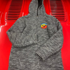 May include: A gray zip-up hoodie with a hood and a small patch on the chest. The patch features a shield design with the word "ABARTH" and a scorpion emblem. The hoodie is made of a textured fabric and has a full-length zipper.