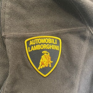 May include: A brown fleece jacket with a full zipper. The jacket features a patch with the words "AUTOMOBILI LAMBORGHINI" in yellow lettering. The patch also includes a yellow bull logo on a black background.
