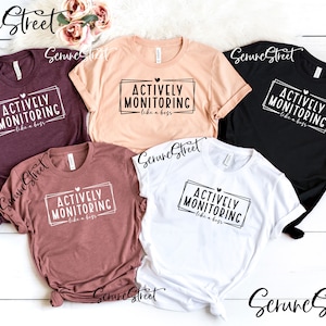 May include: Five t-shirts in different colors with the text "Actively Monitoring like a boss" printed on the front. The shirts are laid out on a white wooden surface.