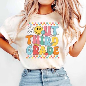 Könnte beinhalten: Ein weißes T-Shirt mit einer bunten Grafik, auf der "Out Third Grade" mit einem Smiley, einem Peace-Zeichen und Schulutensilien steht.