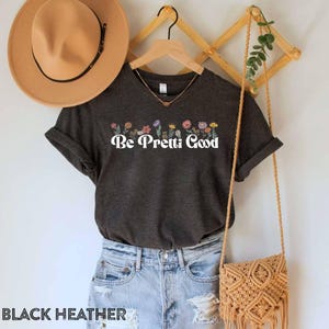 May include: Dark gray t-shirt with the text "Be Pretti Good" in white, above a row of colorful flowers. The shirt is displayed with a brown hat, a gold necklace, and a macrame bag. The words "BLACK HEATHER" are at the bottom.