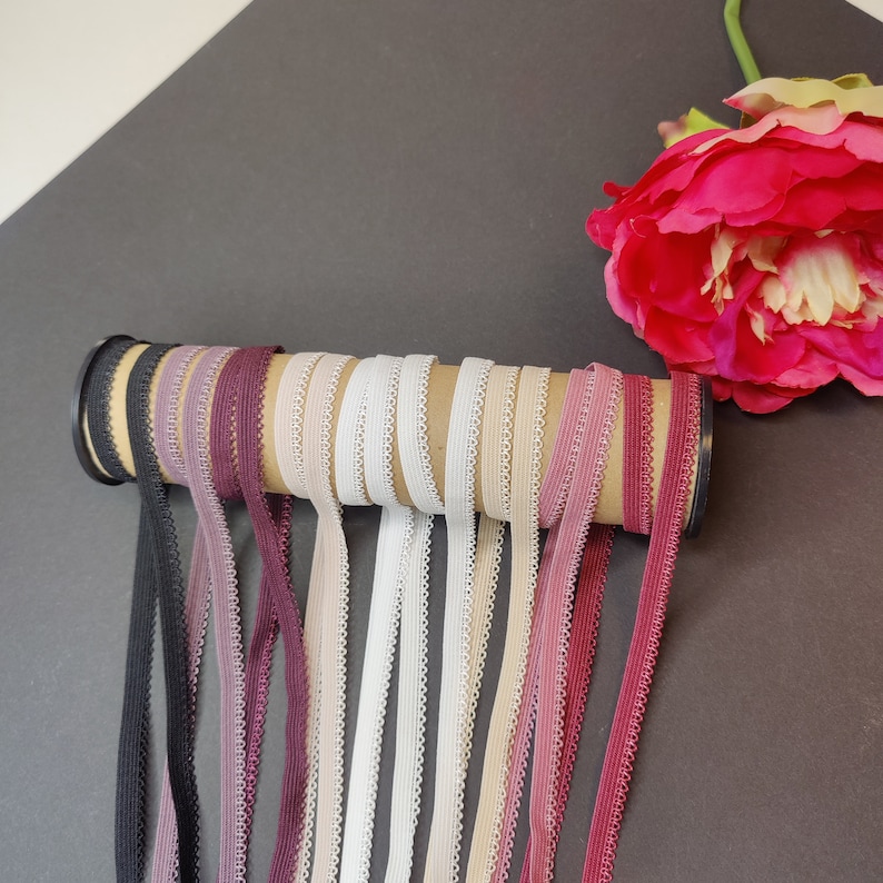 May include: A collection of 10 different colors of elastic trim with a scalloped edge. The trim is rolled up and displayed on a cardboard tube. The trim is available in black, white, beige, pink, and shades of red.