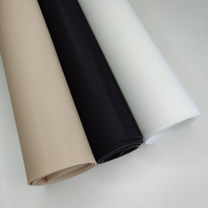 K&ouml;nnte beinhalten: Drei Rollen Stoff in Beige, Schwarz und Wei&szlig;. Der Stoff ist aufgerollt und die Enden sind sichtbar.