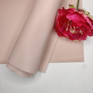 Puede incluir: Un rollo de tela rosa rubor con una sutil textura acanalada. Una vibrante flor de peonía roja y rosa descansa sobre la tela. La tela está cuidadosamente enrollada, mostrando su superficie lisa y color.