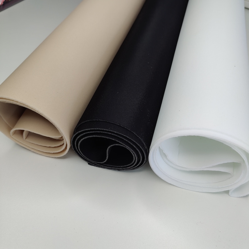 K&ouml;nnte beinhalten: Drei Rollen Stoff in verschiedenen Farben: Beige, Schwarz und Wei&szlig;. Der Stoff ist aufgerollt und die Kanten sind sichtbar.
