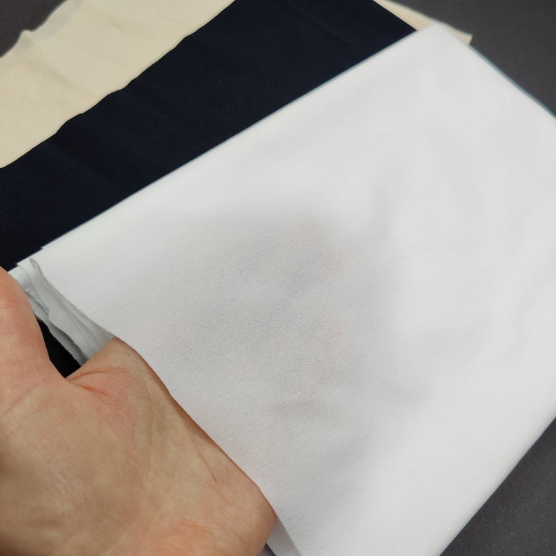 May include: A white fabric swatch, folded in half, with a soft, smooth texture. The fabric is held in a hand, showing its drape and flexibility.