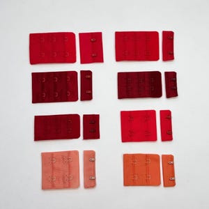 May include: A collection of eight bra hook and eye closure sets in various shades of red, ranging from bright scarlet to deep burgundy and soft coral. Each set consists of a fabric panel with metal hooks and a corresponding fabric strip with metal eyes.