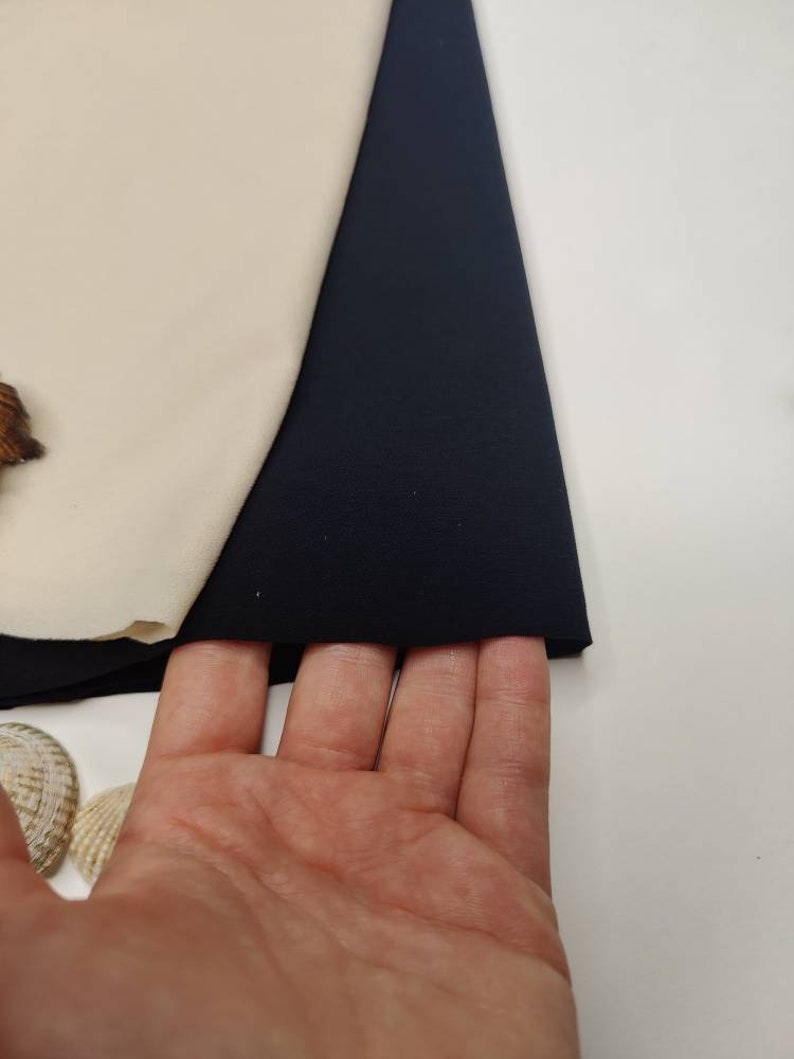 May include: A close-up of a dark blue fabric swatch. The fabric is smooth and has a slight sheen. The swatch is folded in half and is being held by a hand.