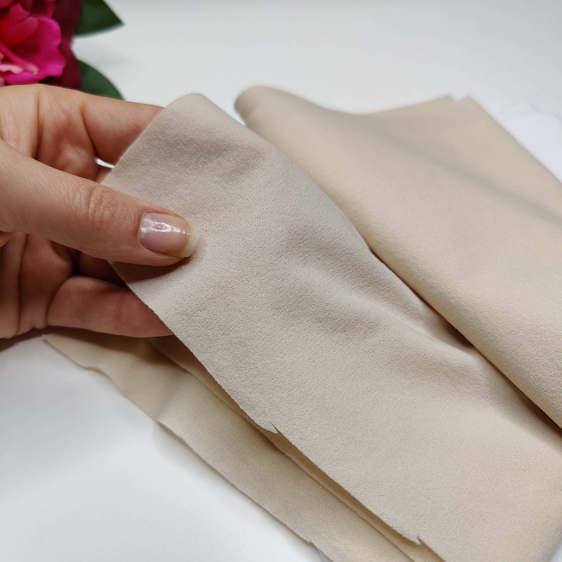 May include: A close-up of a beige microfiber cloth, folded in half, with a hand holding the top layer. The cloth is soft and textured, ideal for cleaning delicate surfaces.