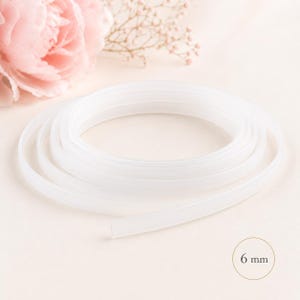 May include: A coiled, translucent white ribbon, 6 mm wide, is the focal point of this image. Soft pink flowers and delicate sprigs of baby's breath are in the background, creating a gentle, romantic aesthetic. The ribbon appears to be made of a flexible material.