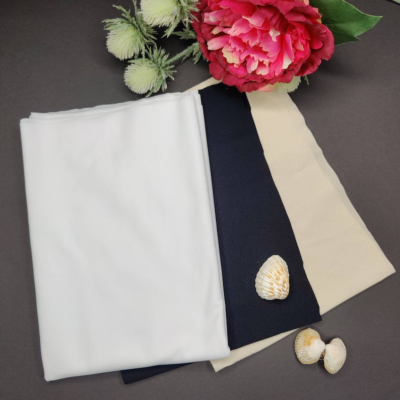May include: Three fabric swatches: white, black, and beige. The swatches are laid out on a dark gray surface.  Two seashells are in the lower right corner of the image.