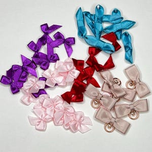 Könnte beinhalten: Sortiment aus Satinband-Schleifen in verschiedenen Farben, darunter rosa, lila, rot und blau. Einige Schleifen haben einen goldenen Ring.