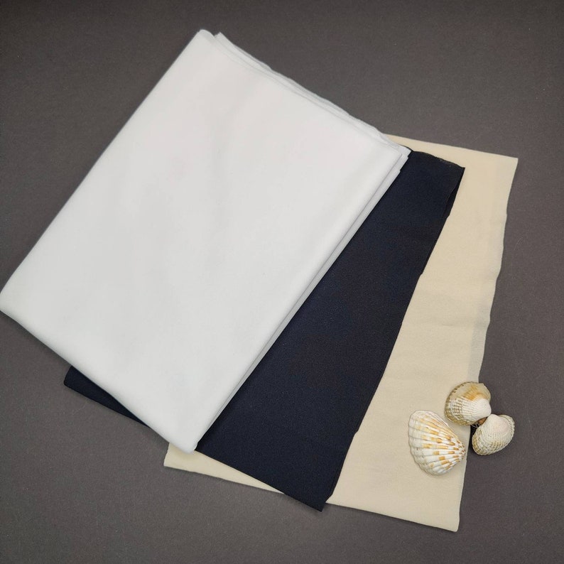 May include: Three fabric swatches, one white, one black, and one beige, are stacked on top of each other. Three seashells are on the beige fabric.