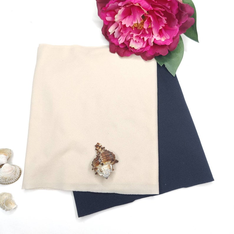 May include: Two fabric swatches, one beige and one navy blue, are laid out on a white surface. A seashell is placed on the beige swatch. A pink flower is in the background.