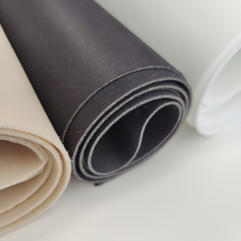 K&ouml;nnte beinhalten: Drei Rollen Stoff in verschiedenen Farben: beige, schwarz und wei&szlig;. Der Stoff ist aufgerollt und die Kanten sind sichtbar.