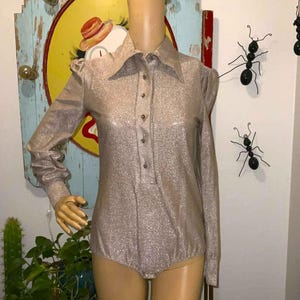 May include: A shimmering, long-sleeved bodysuit in a metallic champagne color. The garment features a collared neckline, button-down front, and a fitted silhouette. The bodysuit is displayed on a mannequin.