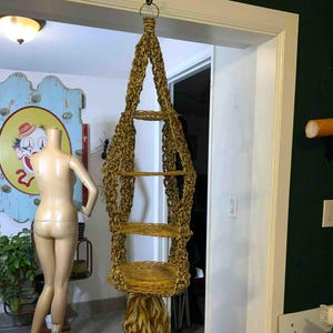 May include: A woven plant hanger with four shelves and a decorative fringe. The macrame-style hanger is made of tan and brown fibers. The plant holder is suspended from a metal hook.
