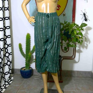 May include: Green, knee-length pants with a drawstring waist and vertical silver stripes. The pants are displayed on a mannequin. A cactus in a blue pot and a vintage sign are in the background.