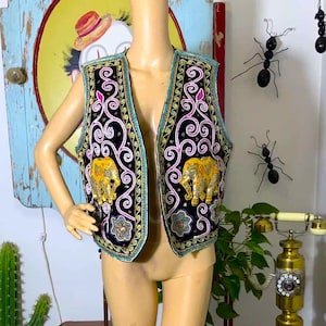 May include: A black sleeveless vest featuring intricate embroidery in pink, white, and gold thread. The garment displays prominent gold elephant motifs on both front panels, accented by decorative floral patterns and a teal blue border trim.