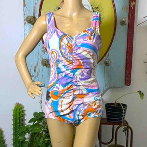 May include: A one-piece swimsuit with a colorful paisley pattern in shades of purple, blue, orange, and white. The swimsuit has a ruched front and wide straps. The mannequin is standing in front of a distressed wooden background.