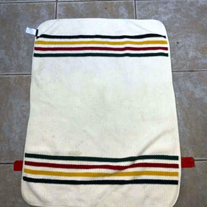 May include: Cream-colored rectangular cushion with horizontal stripes in black, yellow, red, and green. The cushion has a red tab on one side and a white tag on the other. The fabric has a ribbed texture.