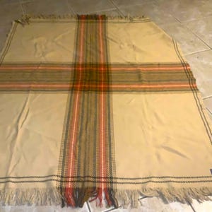 May include: A rectangular beige throw blanket featuring a prominent cross pattern formed by intersecting bands of dark brown, olive green, and bright orange stripes. The blanket is finished with a fringed edge along the top and bottom sides.