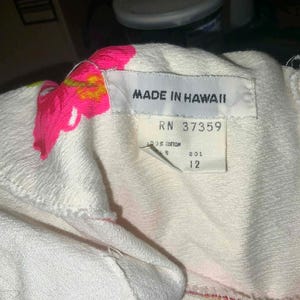 May include: Close-up of a white garment with a tag that reads "MADE IN HAWAII". The garment has pink and yellow floral embroidery. The tag also includes the numbers "RN 37359" and "12".