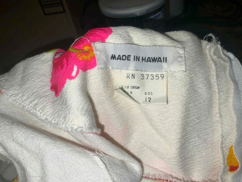 May include: Close-up of a white garment with a tag that reads "MADE IN HAWAII". The garment has pink and yellow floral embroidery. The tag also includes the numbers "RN 37359" and "12".