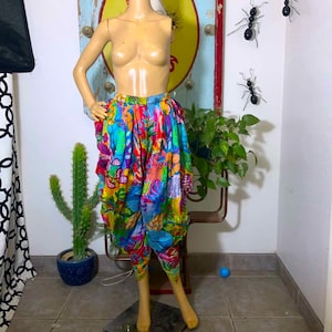 May include: High-waisted harem pants featuring a vibrant, multicolored floral and abstract pattern in shades of blue, yellow, red, and green. The pants have a loose, draped silhouette with gathered fabric at the waist and tapered leg openings.